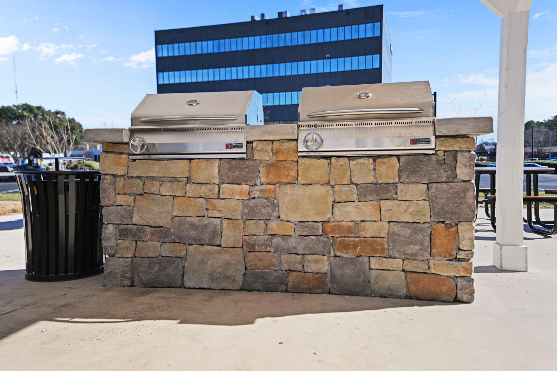 A stone outdoor grill island with two stainless steel grills set on a concrete patio in front of a modern office building.