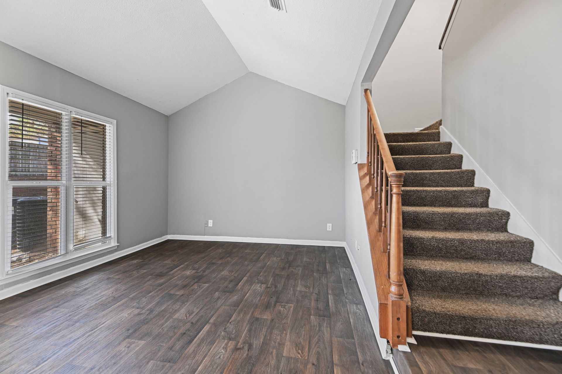 A room with dark wood flooring, light grey walls, a large window, and carpeted stairs with a wooden railing.