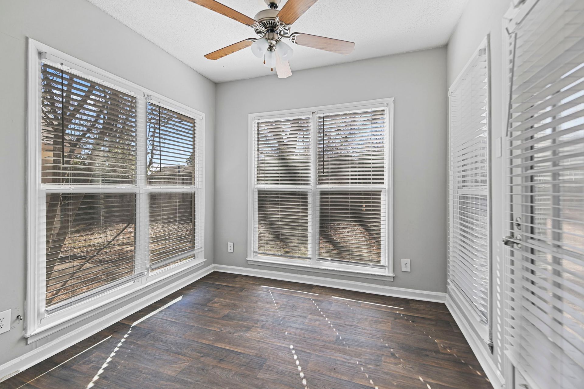 A bright, empty sunroom with light gray walls, dark wood-look flooring, large windows with white blinds, and a ceiling fan.