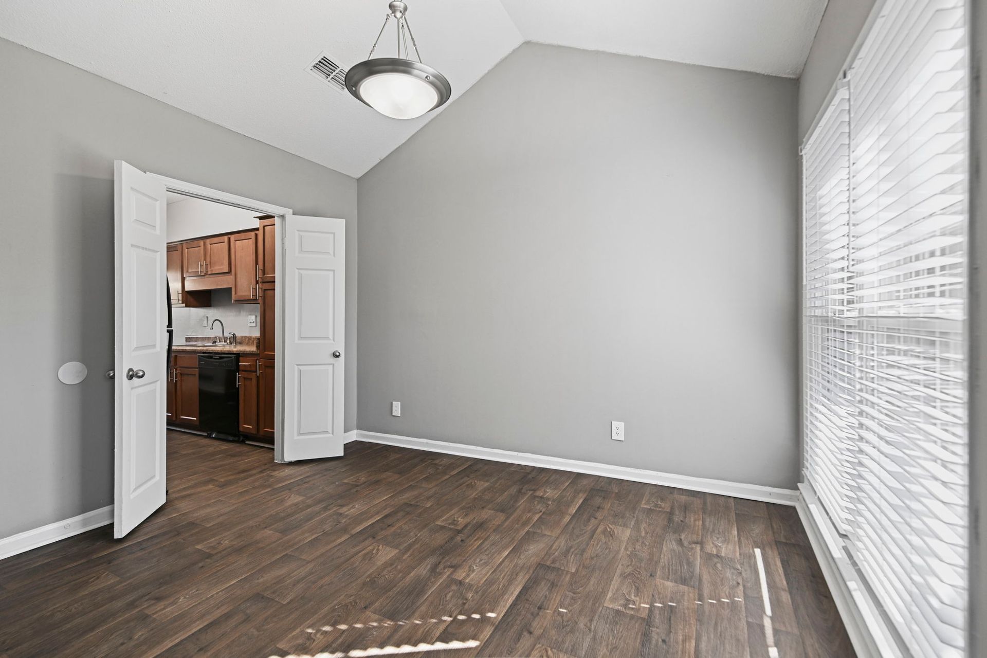 A bright room with light gray walls, dark wood-look floors, a vaulted ceiling, and a doorway leading to a kitchen.