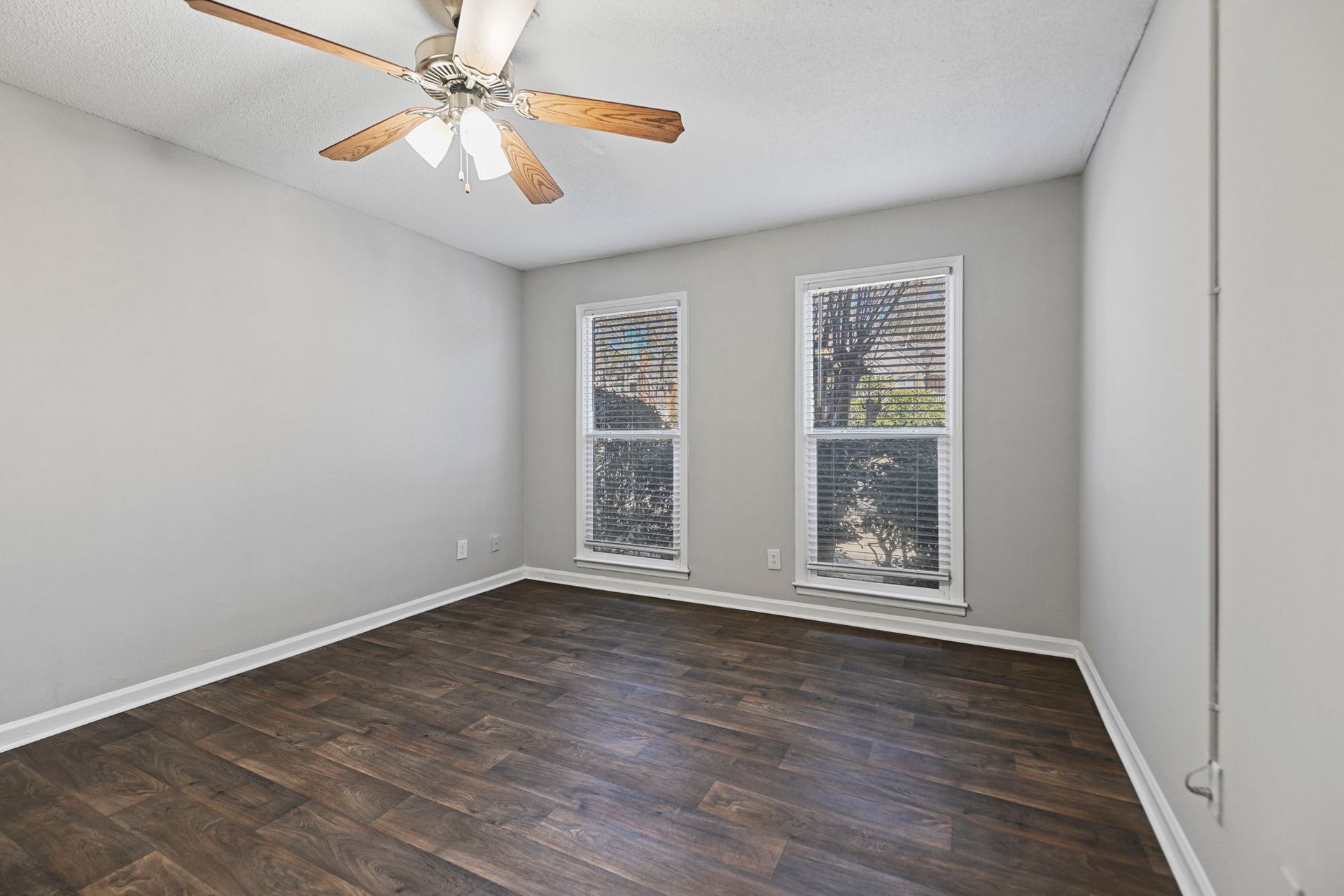 An empty room with light grey walls, dark wood floors, a ceiling fan, and two windows showing an outdoor view.