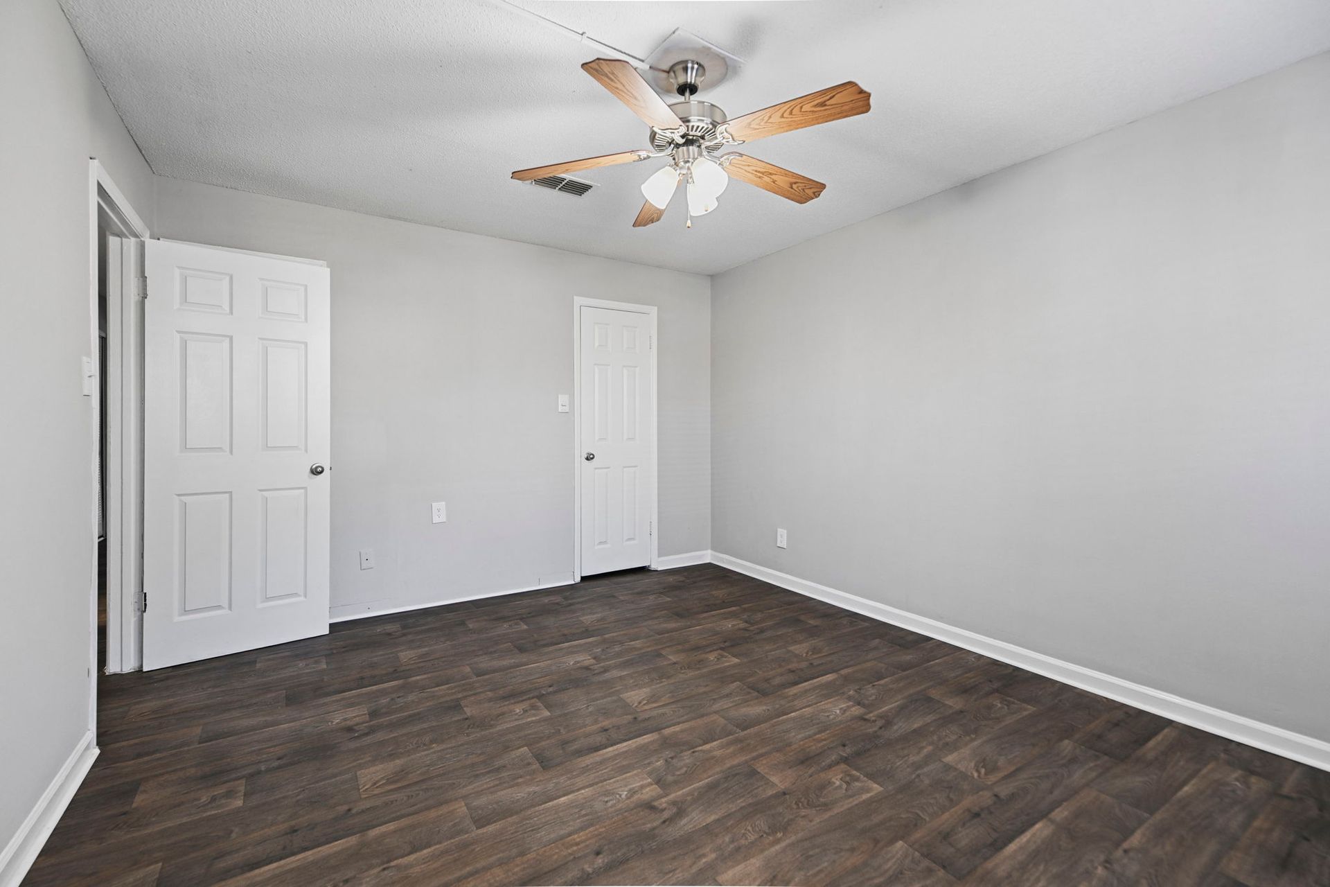 An empty room with light gray walls, dark wood-look floors, a white door, a closet, and a ceiling fan with lights.
