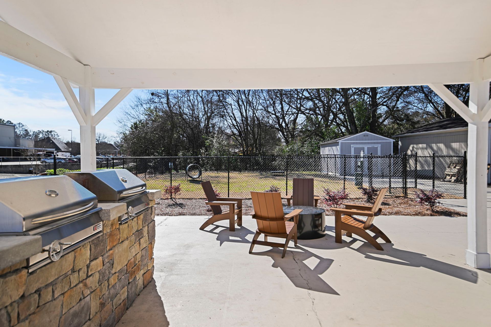 A covered patio features a stone grill station and several wooden Adirondack chairs arranged around a small fire pit.