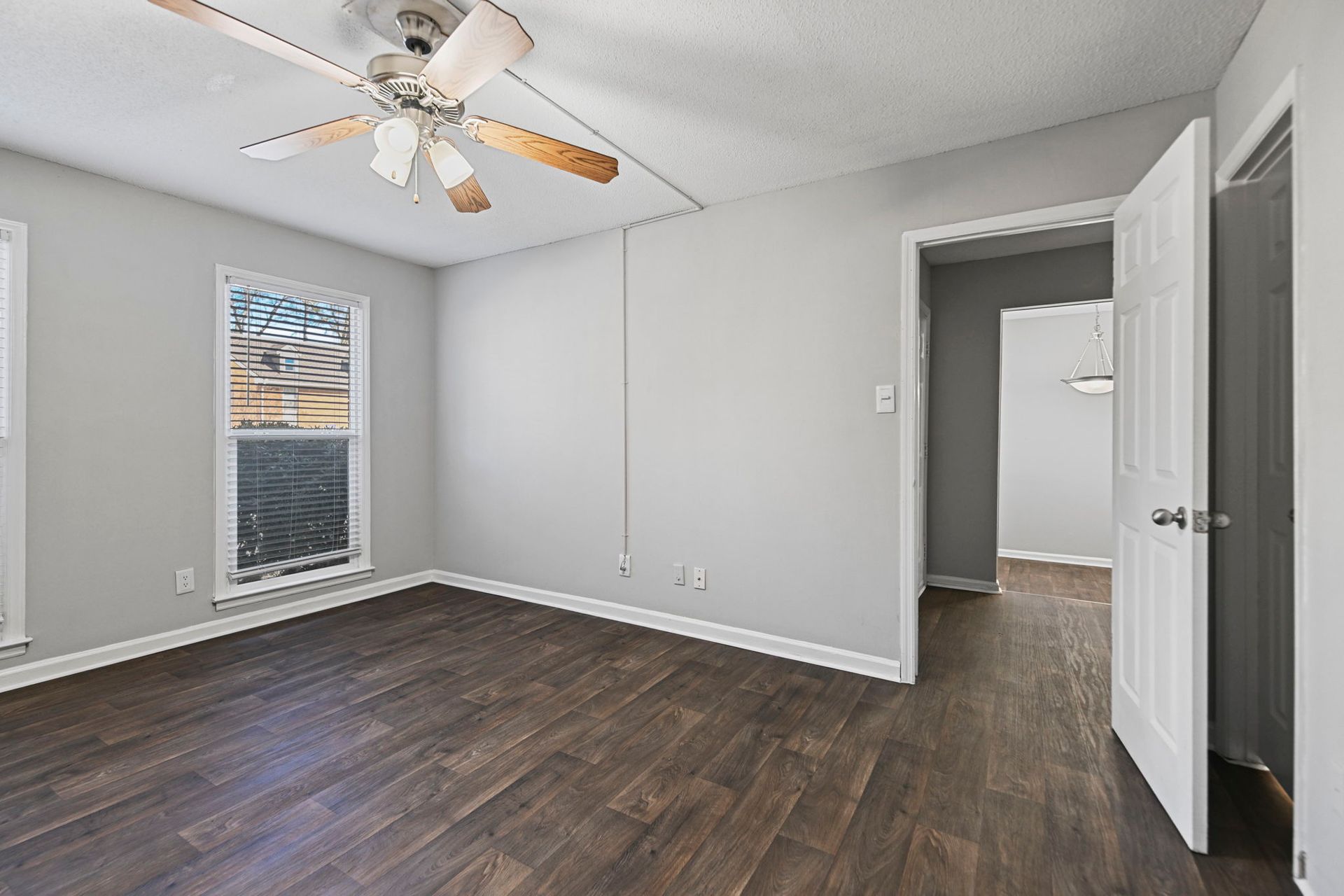 An empty room with light gray walls, dark wood-look floors, a ceiling fan, a window, and an open door to a hallway.