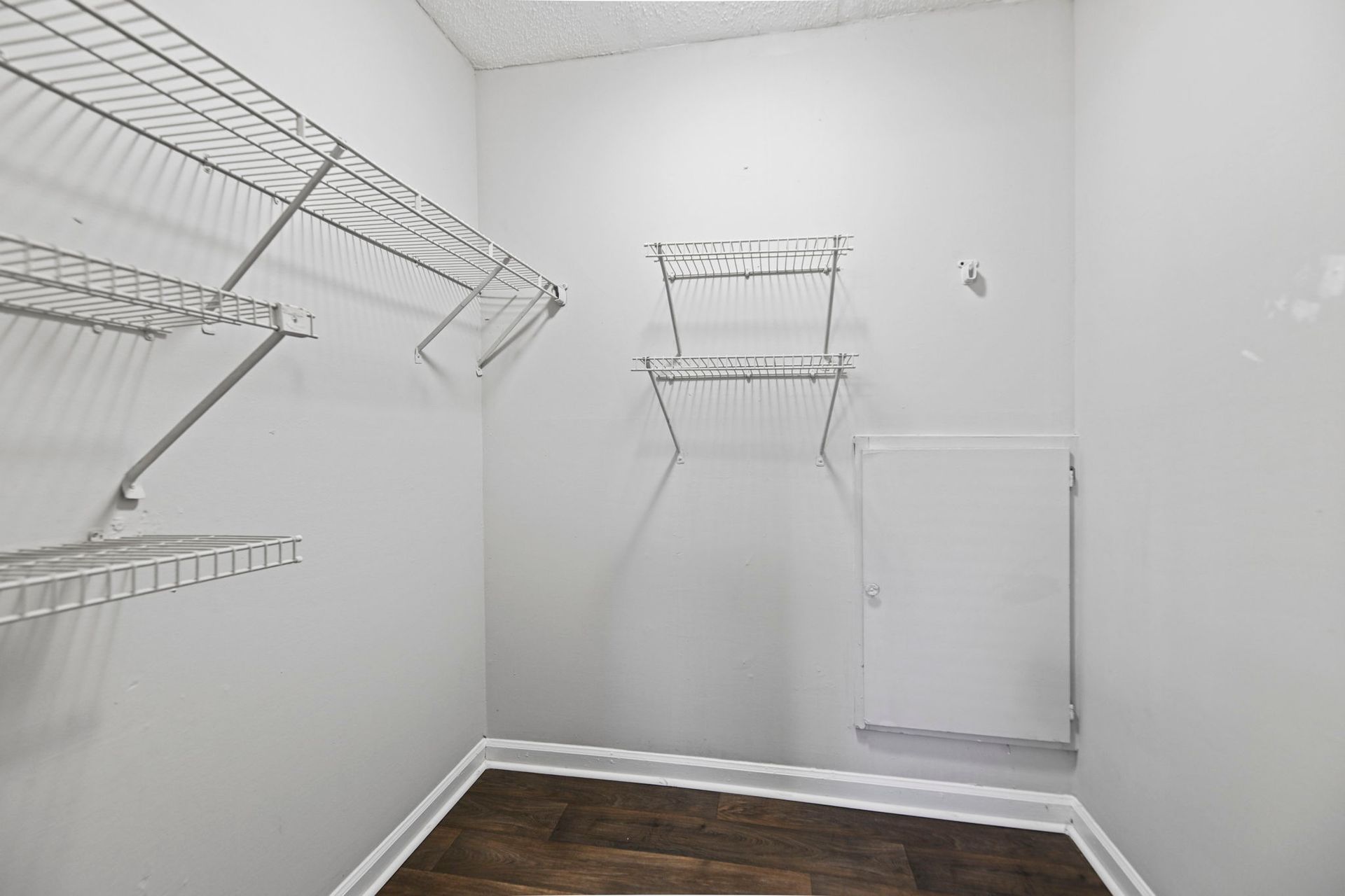 An empty walk-in closet with white walls, dark wood-look flooring, wire shelving, and a small white access panel.