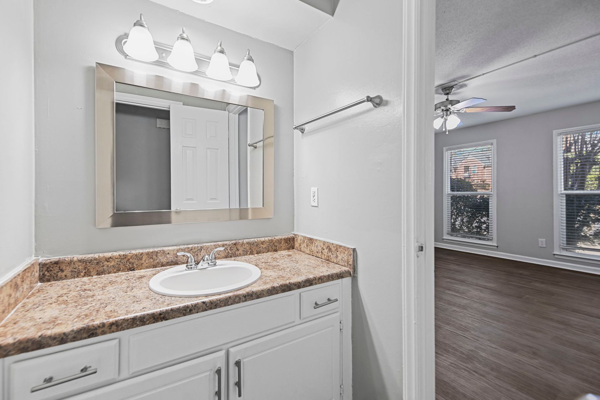 A bathroom vanity with a speckled countertop and white cabinets, adjacent to a room with dark wood floors and windows.