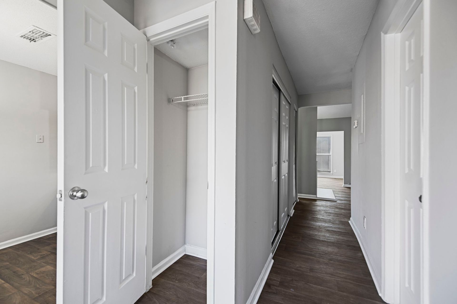 A hallway with white walls and dark wood flooring, featuring a partially open door, an open closet, and a distant doorway.