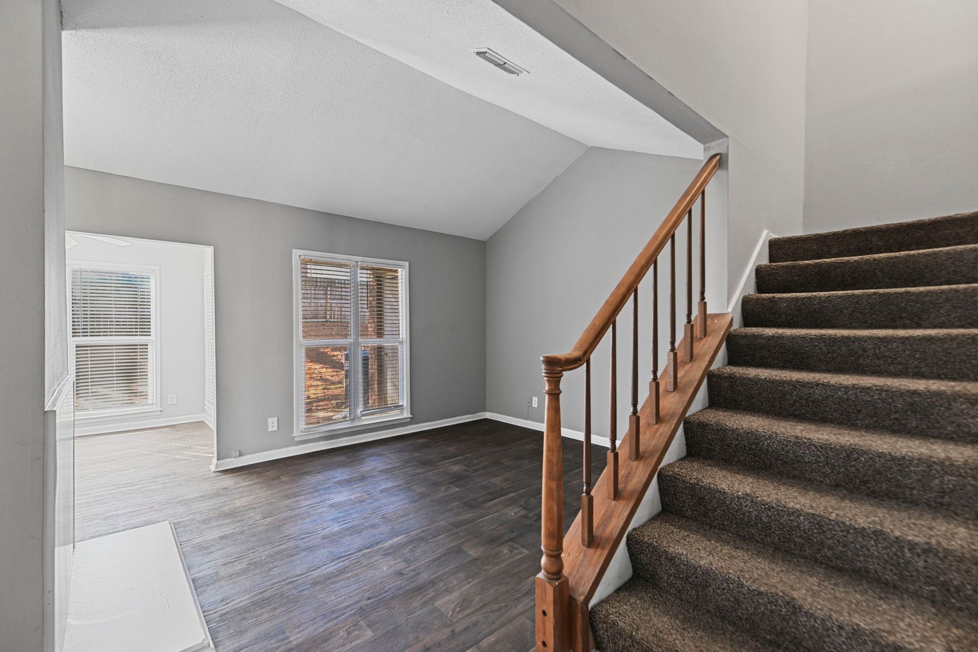 An interior view showing a staircase with brown carpeting, wooden railings, grey walls, and wood-look flooring.
