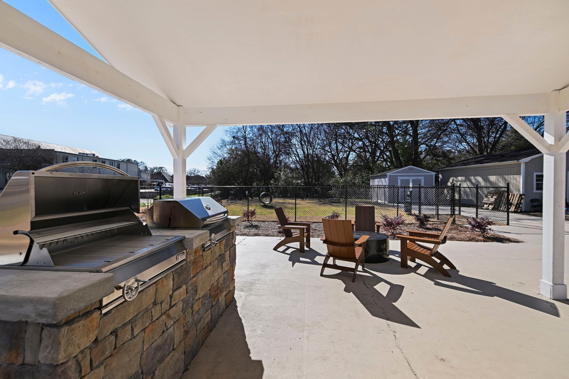 Outdoor patio area with a stone-base grill, wooden chairs, and a fire pit under a white pergola on a sunny day.