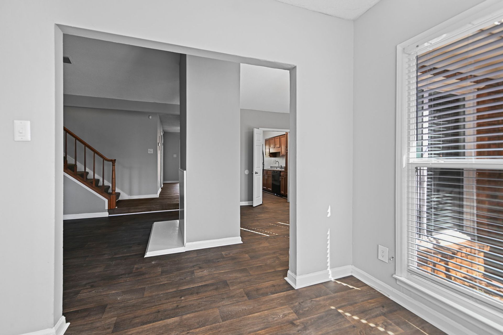 An interior view of a room with light grey walls, dark wood floors, and a window with horizontal blinds.