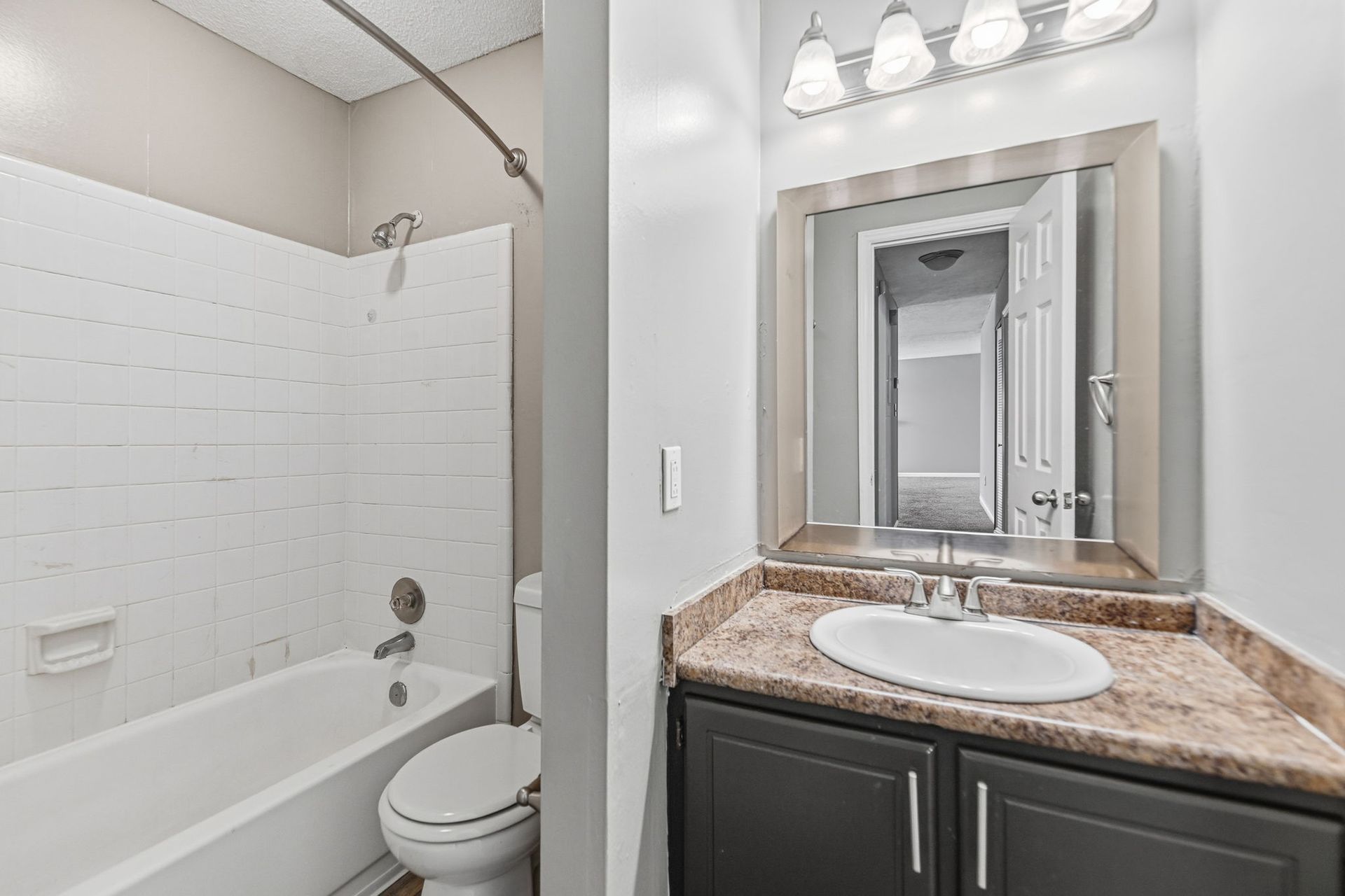 A bright bathroom featuring a white tub, a toilet, a vanity with a granite countertop, and a large mirror with lighting.