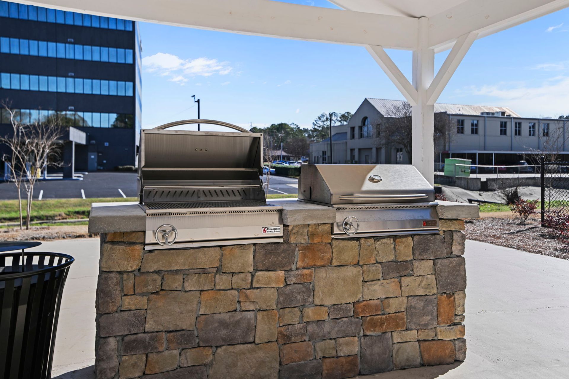 An outdoor stone grilling station with two stainless steel grills under a white pergola on a sunny day.