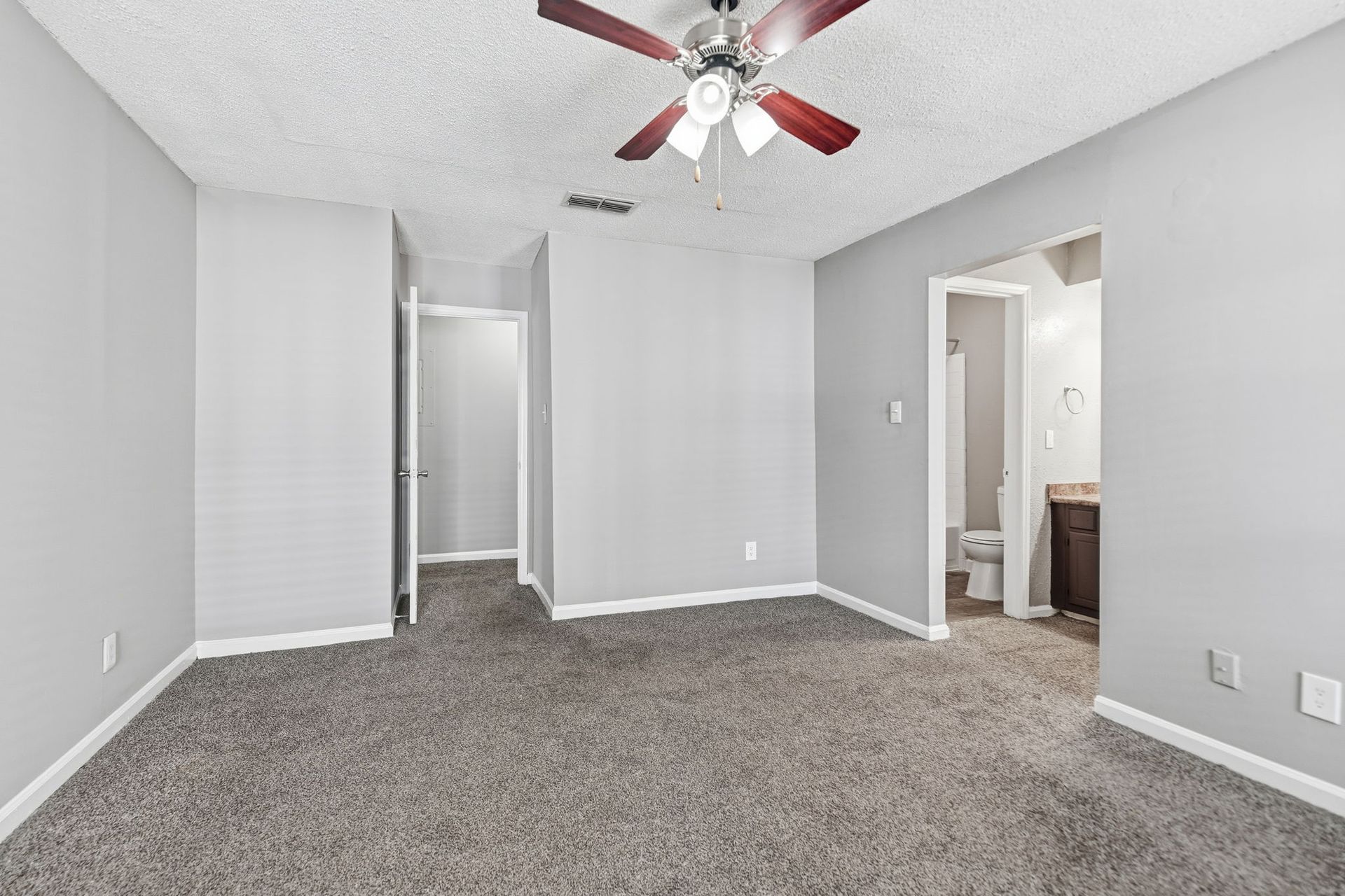 Empty bedroom with gray carpet, light gray walls, a ceiling fan, and two doorways leading to a bathroom and hallway.