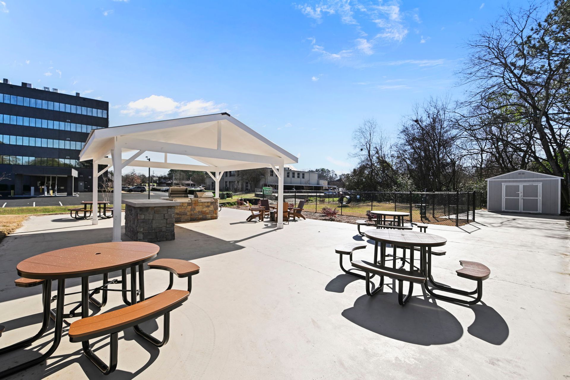 Outdoor patio with picnic tables, a covered grilling station, and a small shed under a clear blue sky.