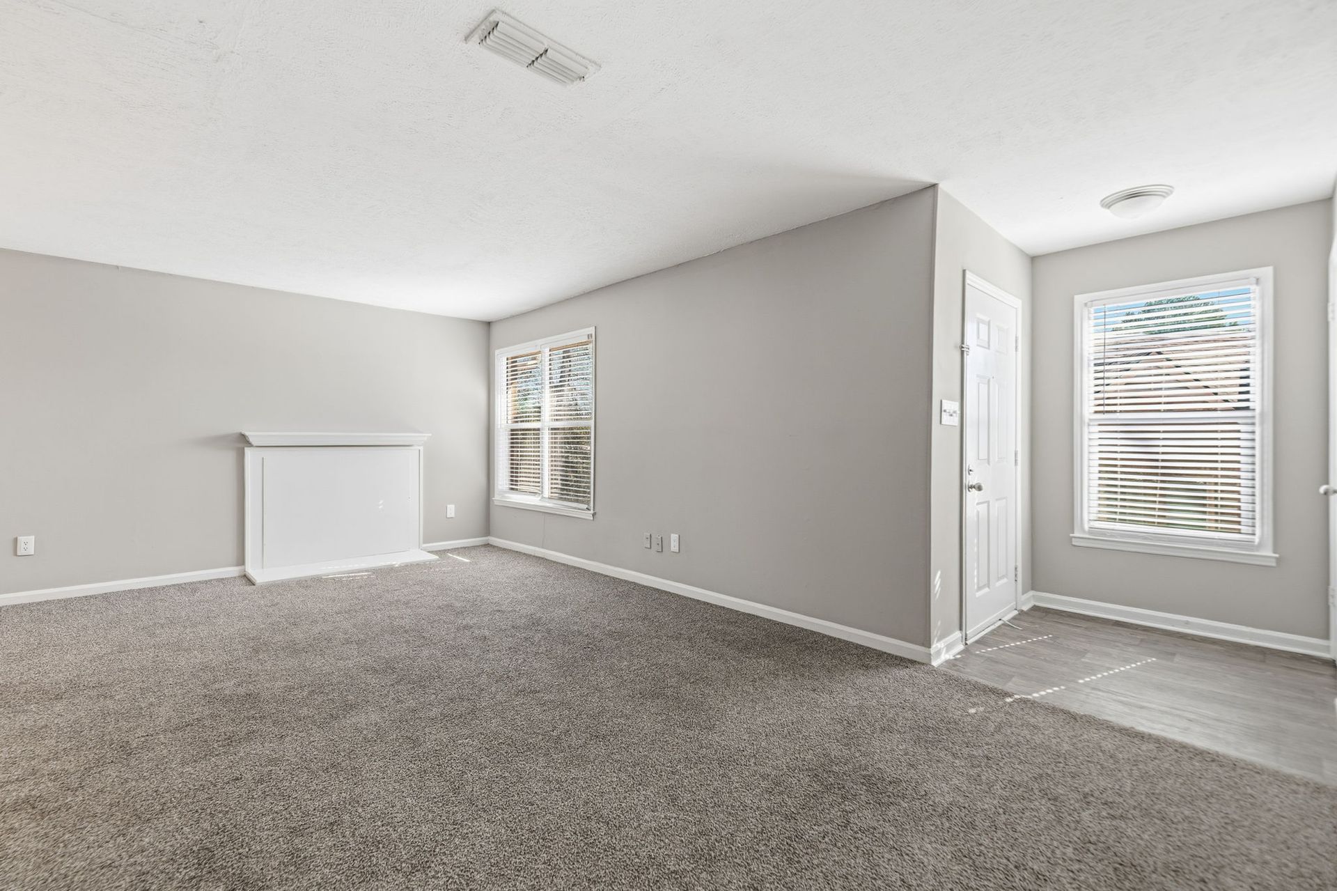 An empty living room with light gray walls, plush gray carpet, a white fireplace, and a window with blinds.