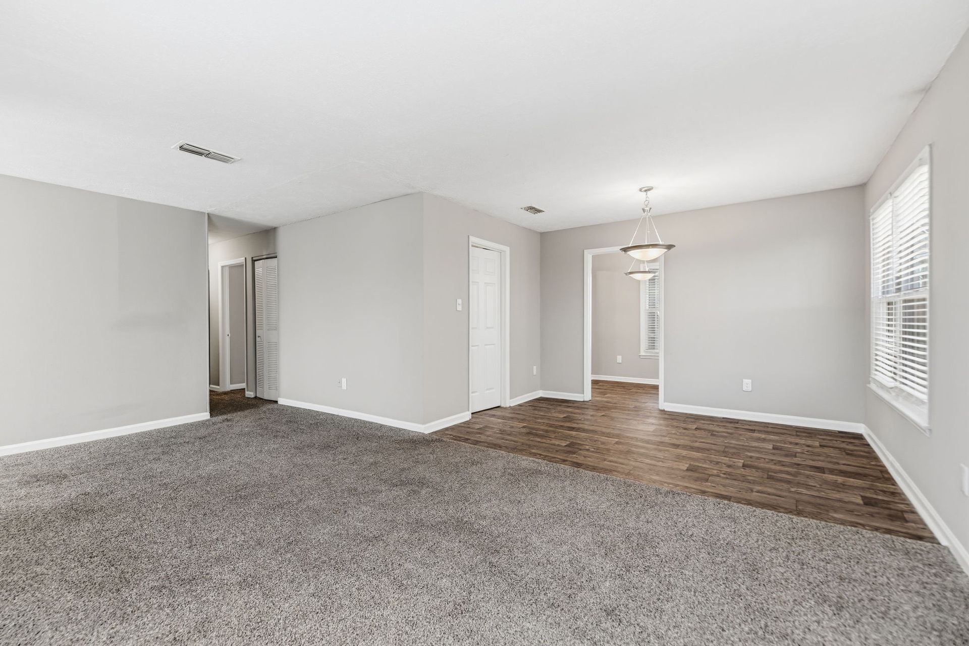 Empty room with gray carpet and wood-look flooring, light gray walls, and a transition to a hallway and kitchen area.