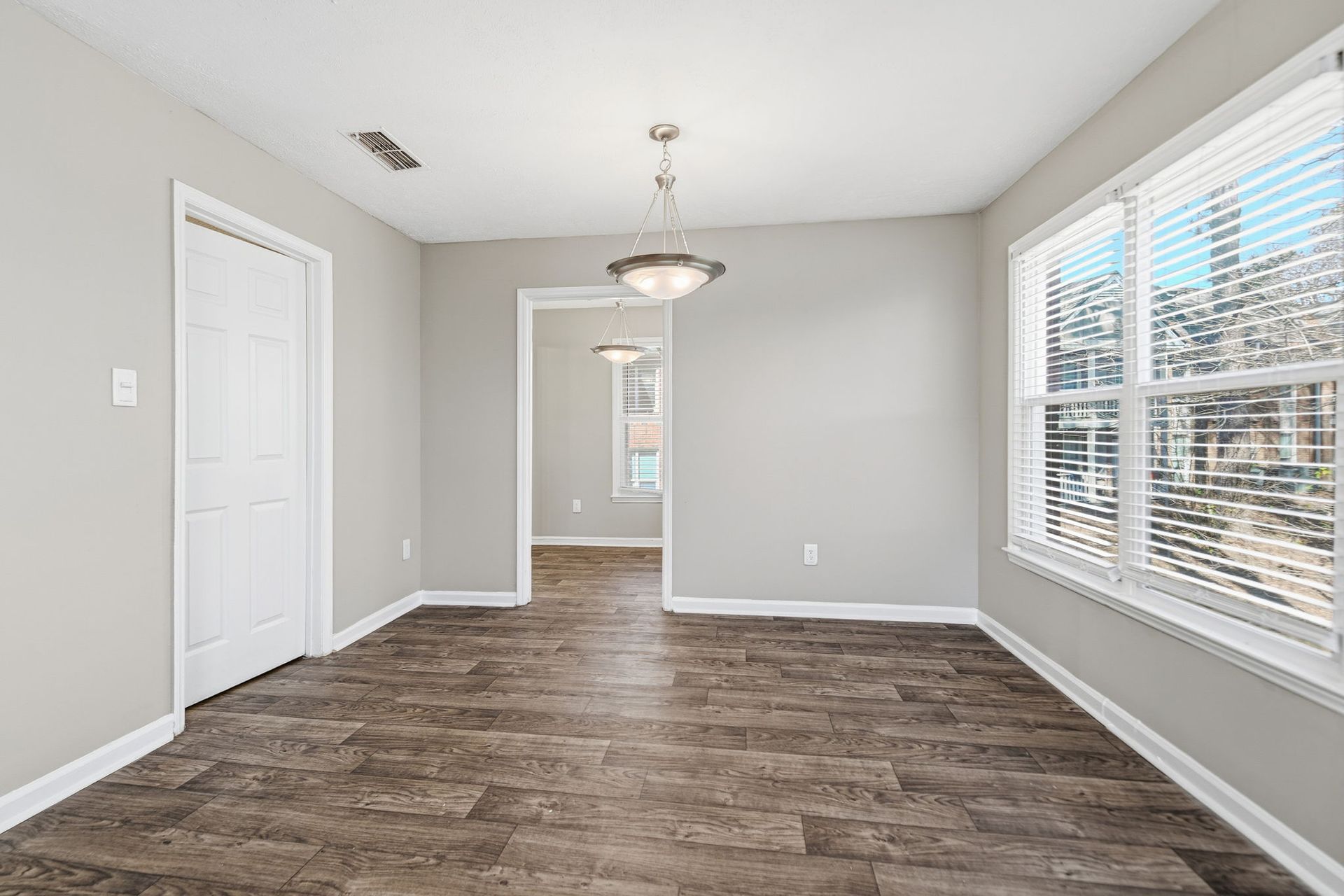 An empty room with light gray walls, dark wood-look floors, a white door, and a modern chandelier.