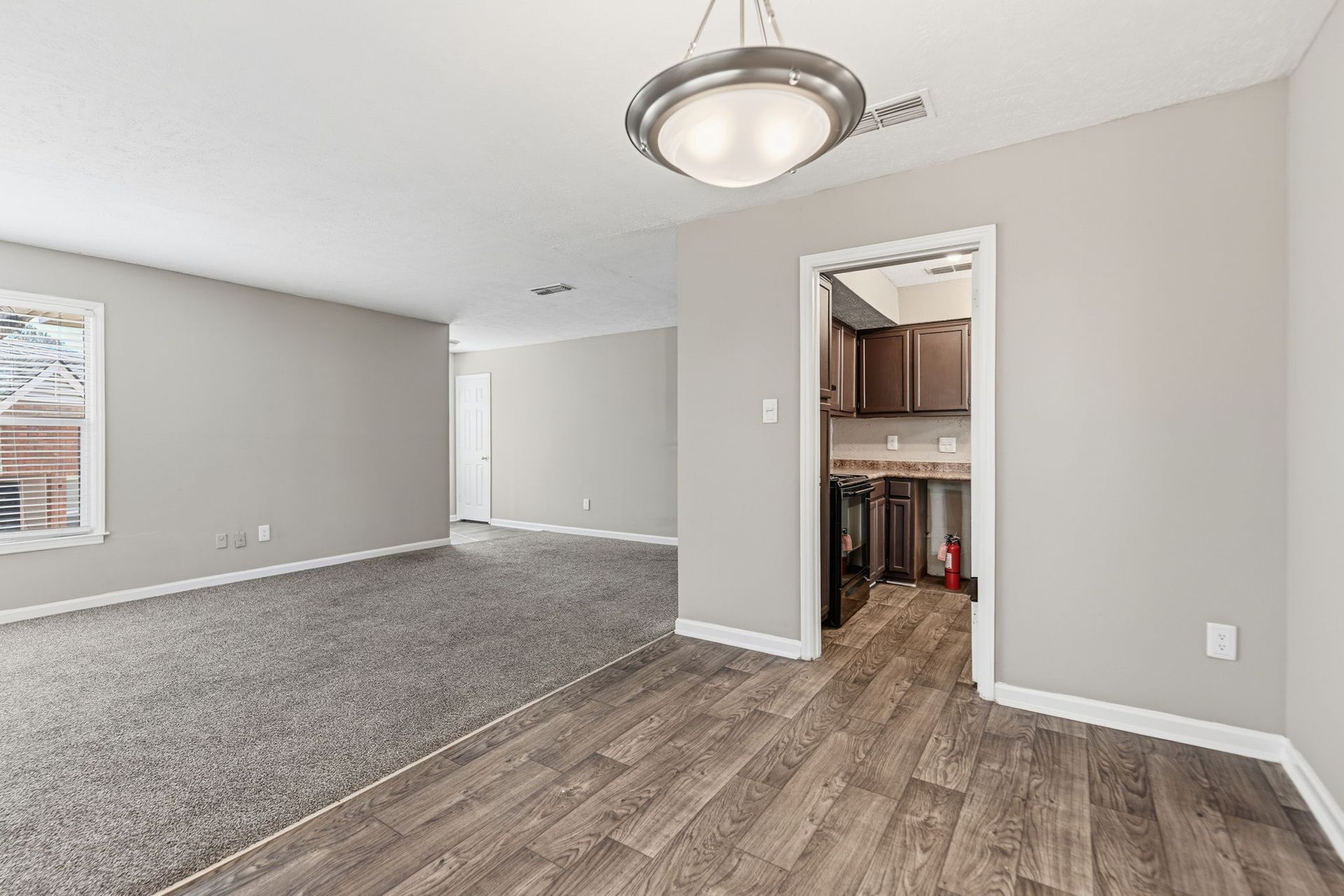 A living room with gray carpet leads to a dining area with wood-look flooring and a view into a dark-toned kitchen.