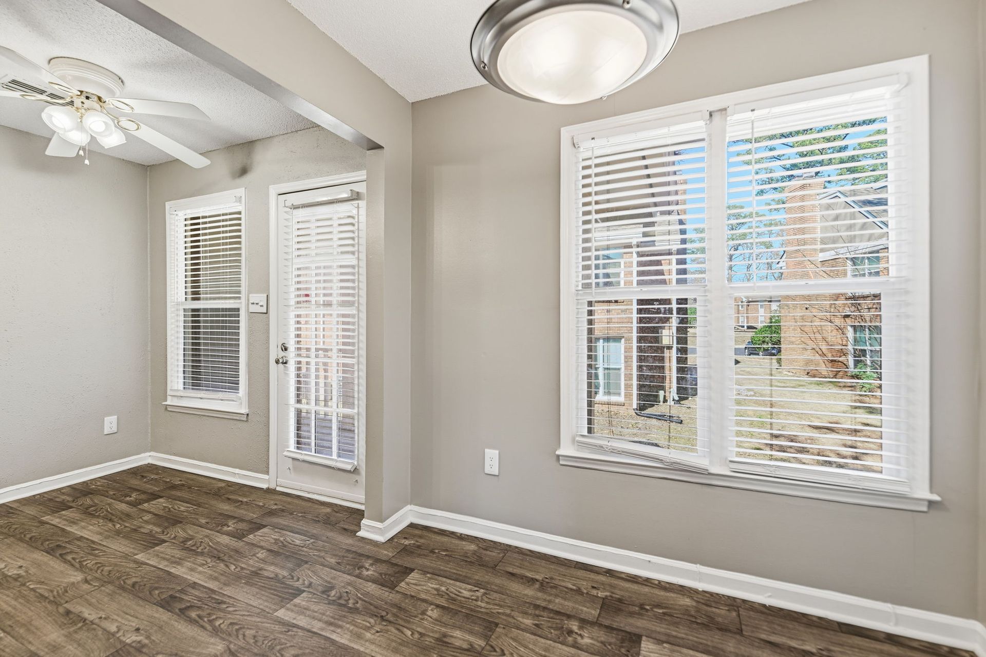 A bright room with light walls and wood-patterned flooring, featuring a ceiling fan, sliding door, and large window.