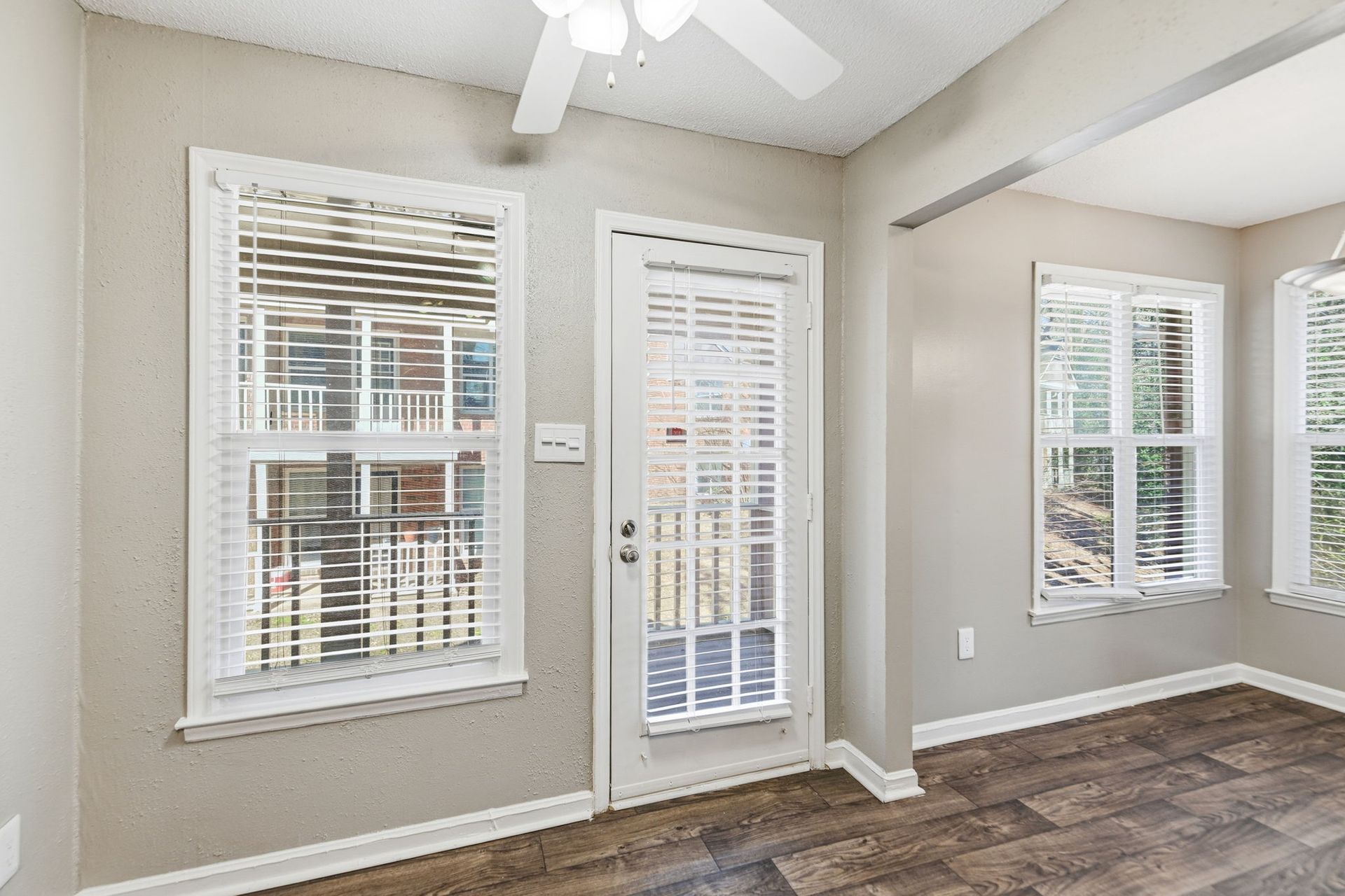 A bright, neutral-toned room featuring two windows and a glass-paneled door, all with white blinds, and dark wood floors.