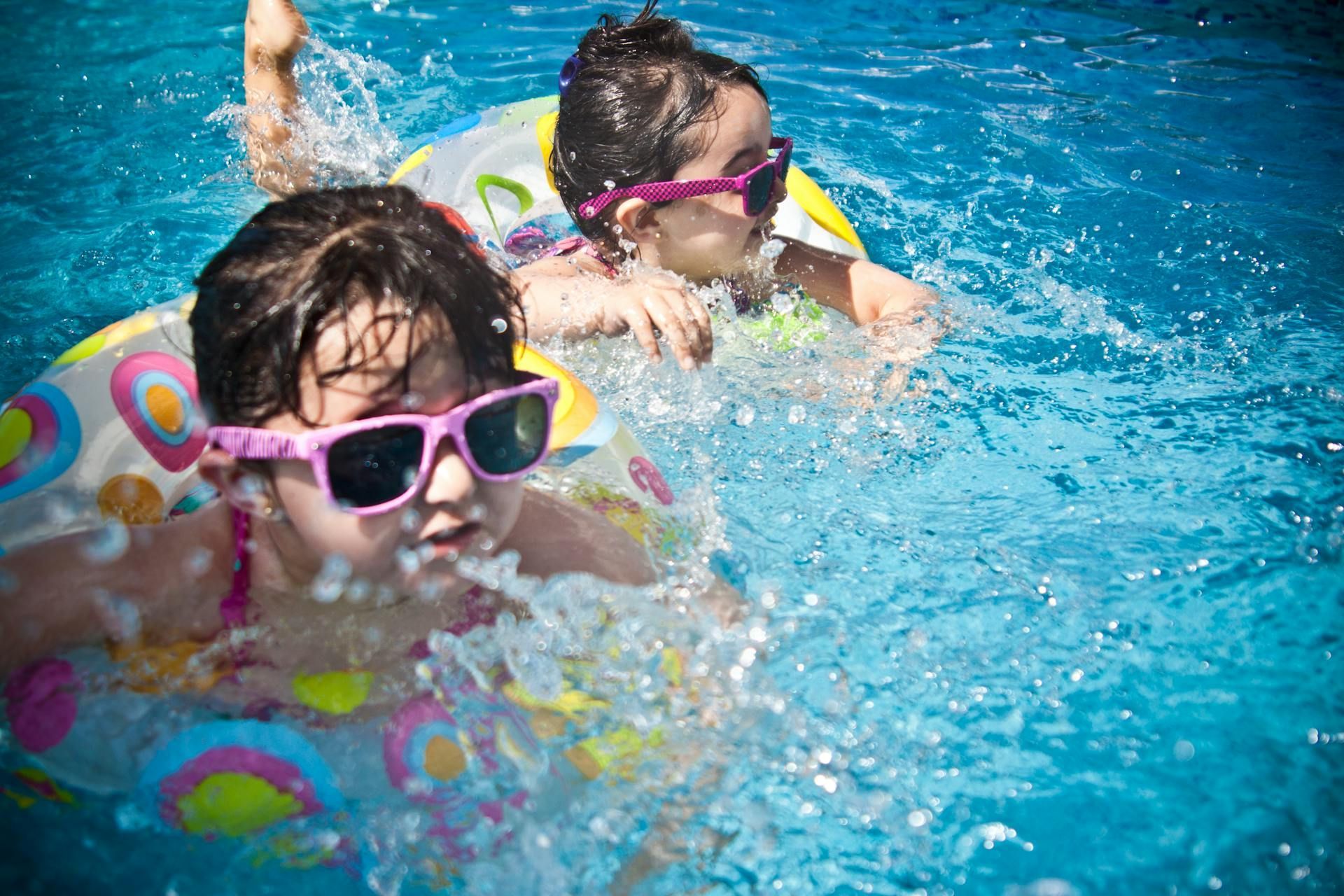 Two children wearing pink sunglasses swim in a pool with colorful inflatable rings.