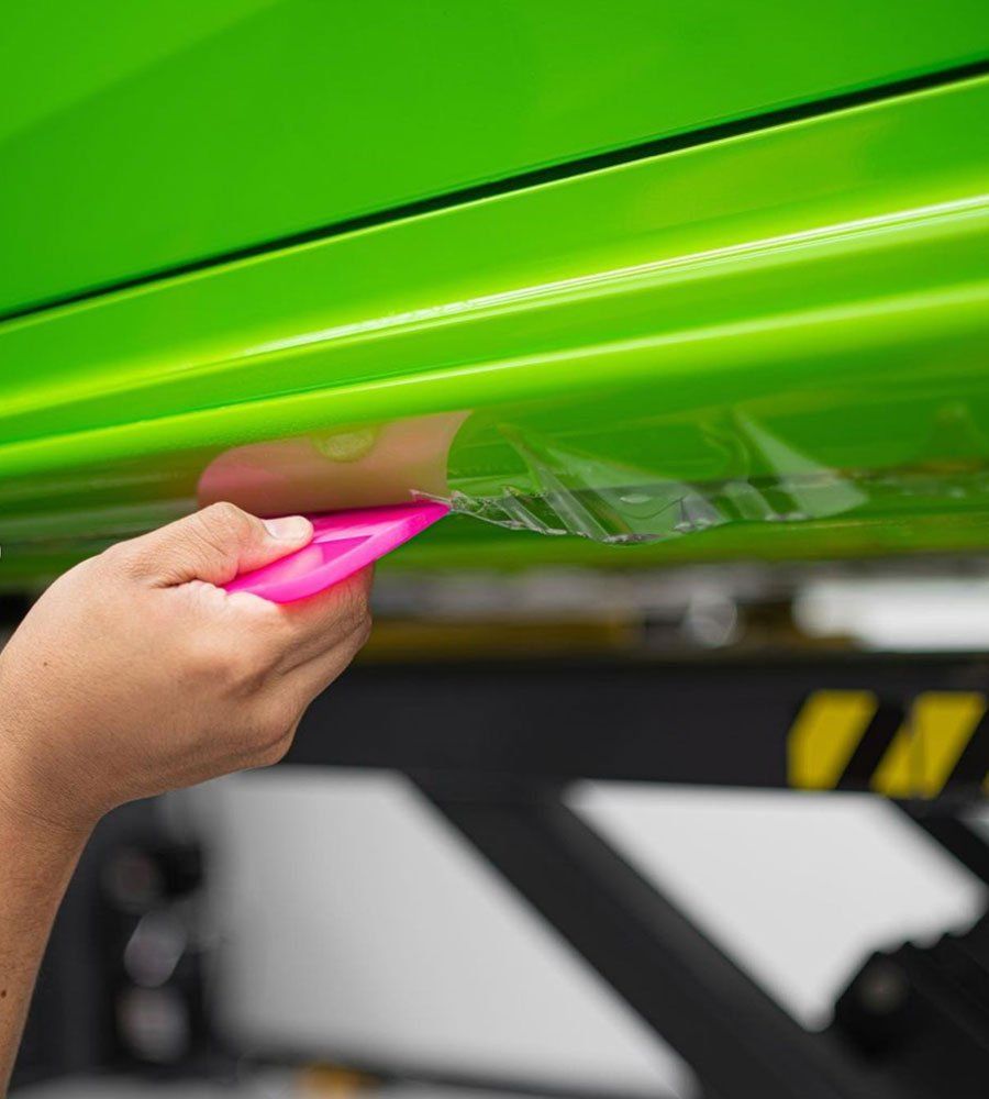 A person is wrapping a green car with a pink tool.