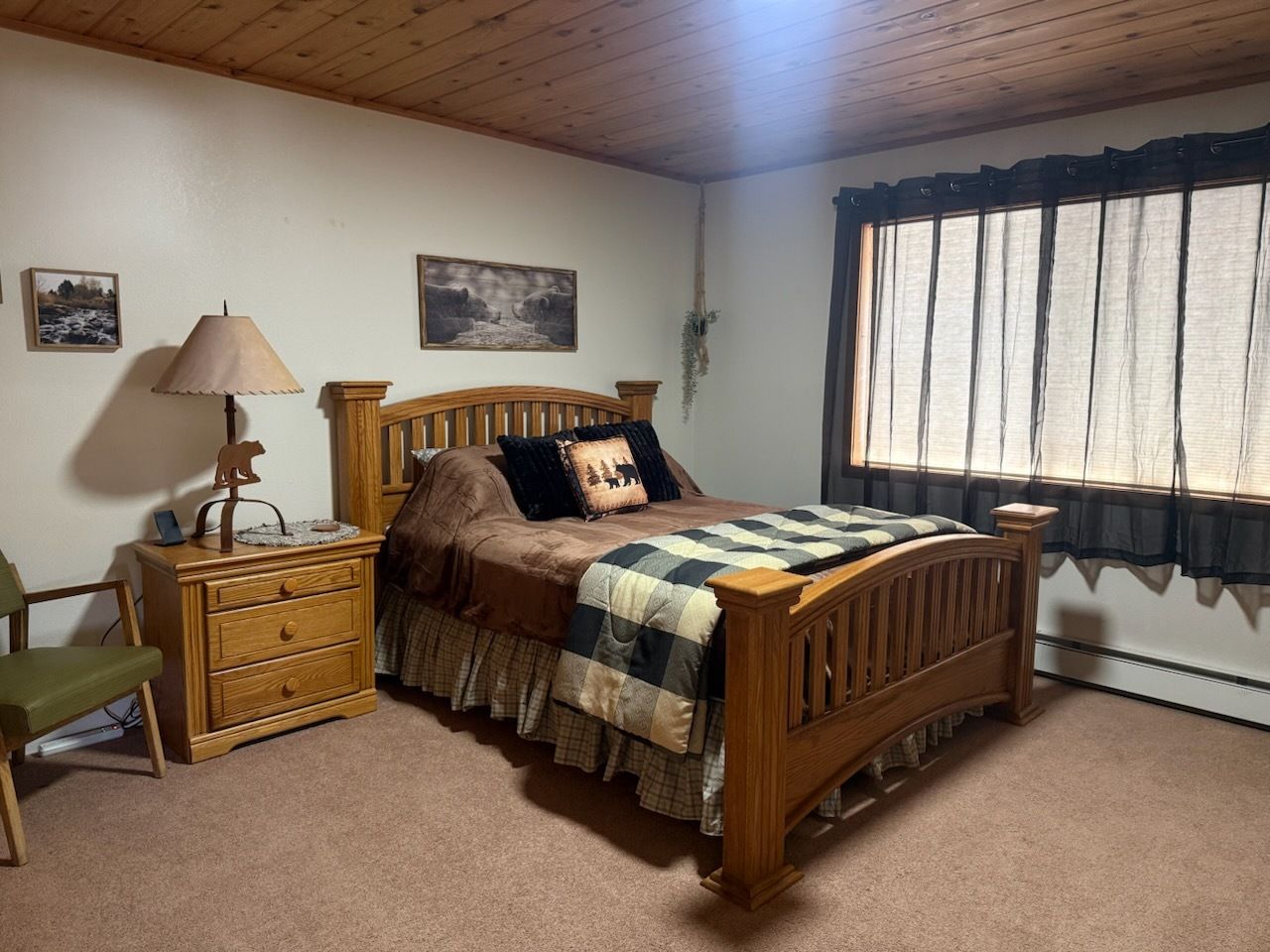 Cozy bedroom with wooden bed, plaid quilt, bedside lamp, and large window with curtains.