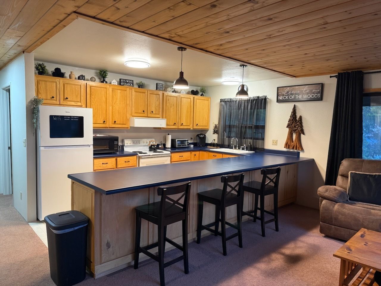 Cozy kitchen and living area with wood cabinets, black island stools, and a brown sofa under a sloped wooden ceiling