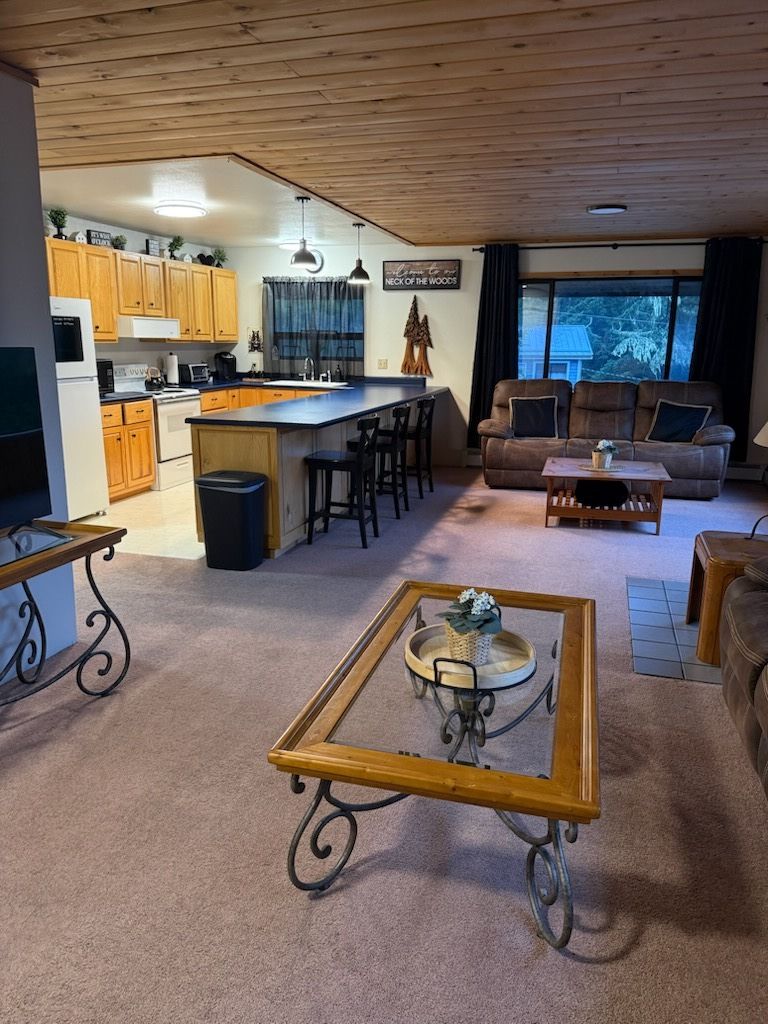 Spacious living room with open kitchen, wood ceiling, dark sofa, bar stools, and glass coffee table.