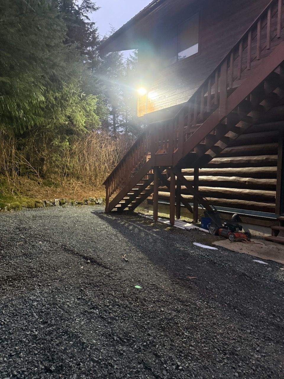 Gravel driveway beside a log cabin staircase in bright sunlight, with trees and a rocky hillside nearby