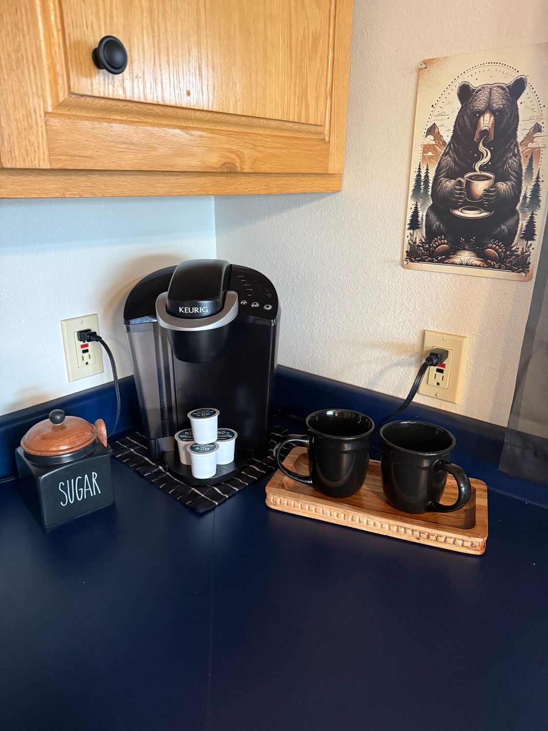 Coffee station on a navy countertop with a Keurig, mugs, sugar container, and wall decor