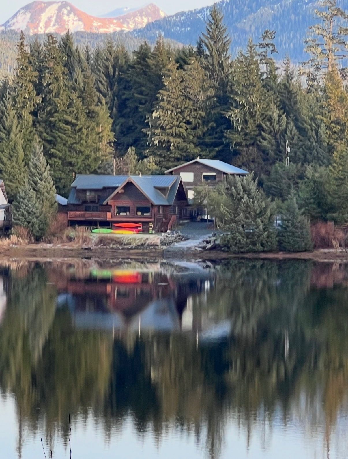Lakeside cabins reflected in a calm lake, framed by pine trees and mountain peaks.