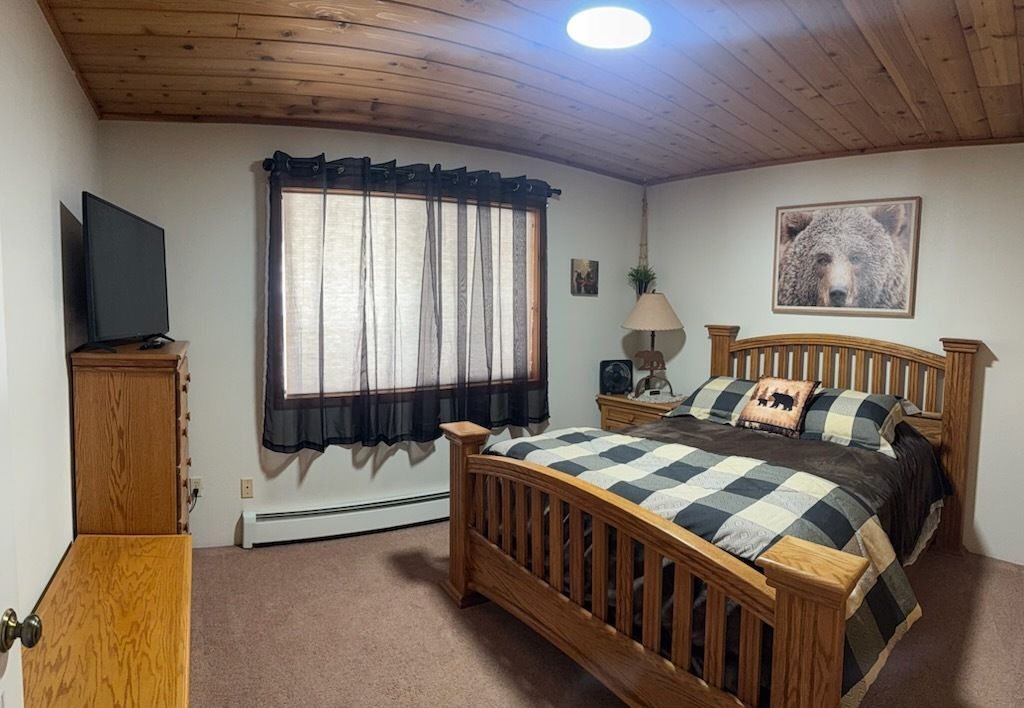 Cozy bedroom with wooden bed, plaid bedding, TV, and curtained window under a wood-paneled ceiling