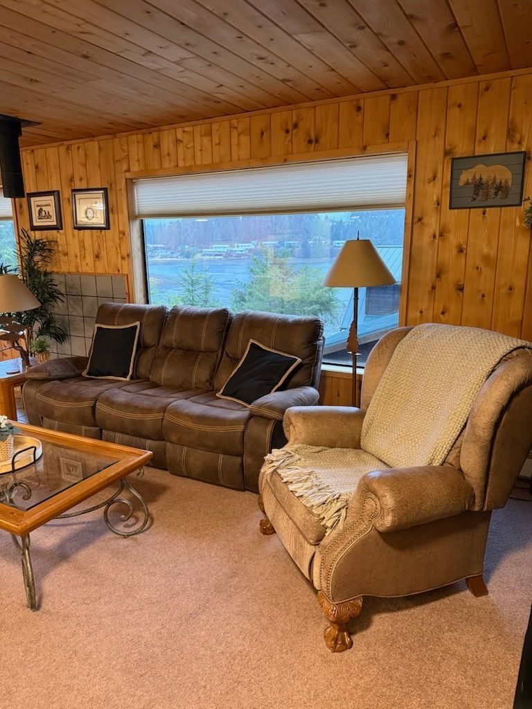 Cozy wood-paneled living room with brown sofa, beige chair, coffee table, lamp, and large window view