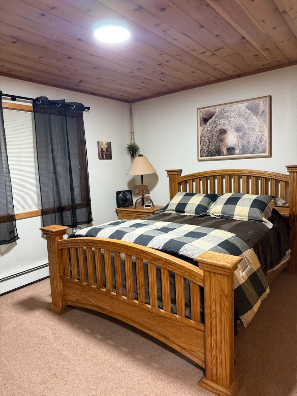 Cozy bedroom with a wooden bed, plaid quilt, lamp, window curtains, and wood-paneled ceiling.