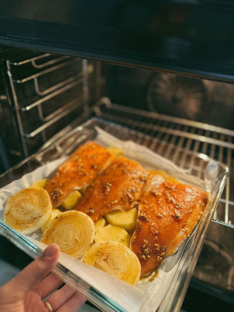 Hand placing a tray of roasted chicken, sliced potatoes, and rolls into an oven