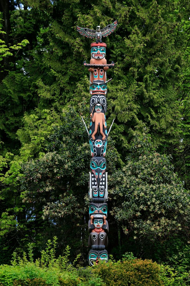 Tall carved totem pole with colorful faces standing in dense green forest.