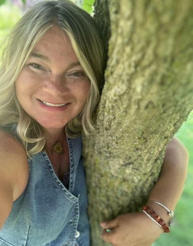 Woman with blonde hair smiles, hugging a tree trunk, wearing a denim vest in a green outdoor setting.