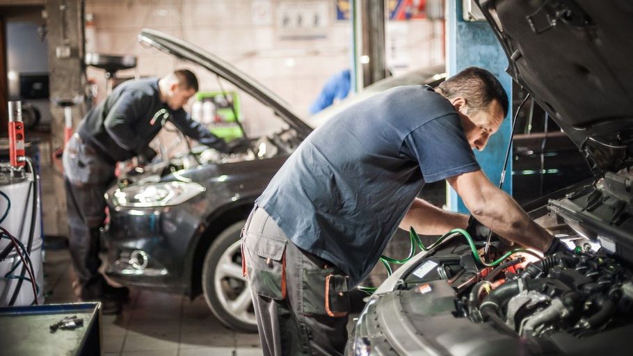 Two mechanics work on cars in an auto repair shop, one inspecting an engine with the hood open.