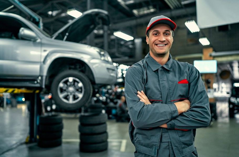 Mechanic smiling with arms crossed in an auto repair shop beside a raised silver truck