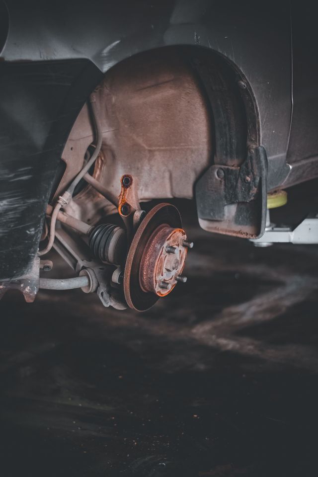 Car’s rear wheel removed, exposing rusty brake rotor and suspension in a dim garage.