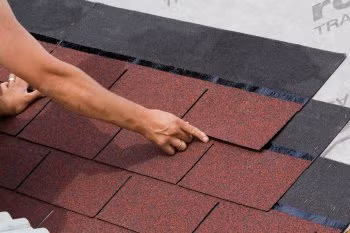 Hand placing red roof shingles on a sloped roof with black underlayment