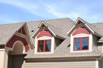 Suburban house with gray roof, red dormer windows, and beige garage under a blue sky