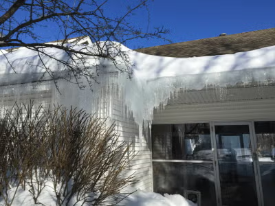 Snow and large icicles hanging from a house roof above a window and shrubs