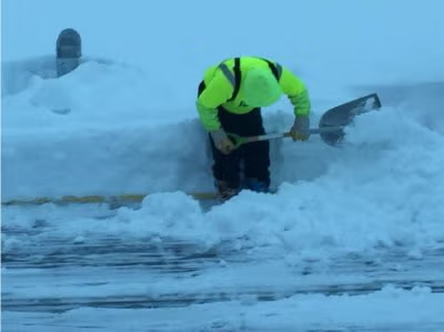 Worker in a neon jacket shoveling snow beside a snowbank in a snowy outdoor area