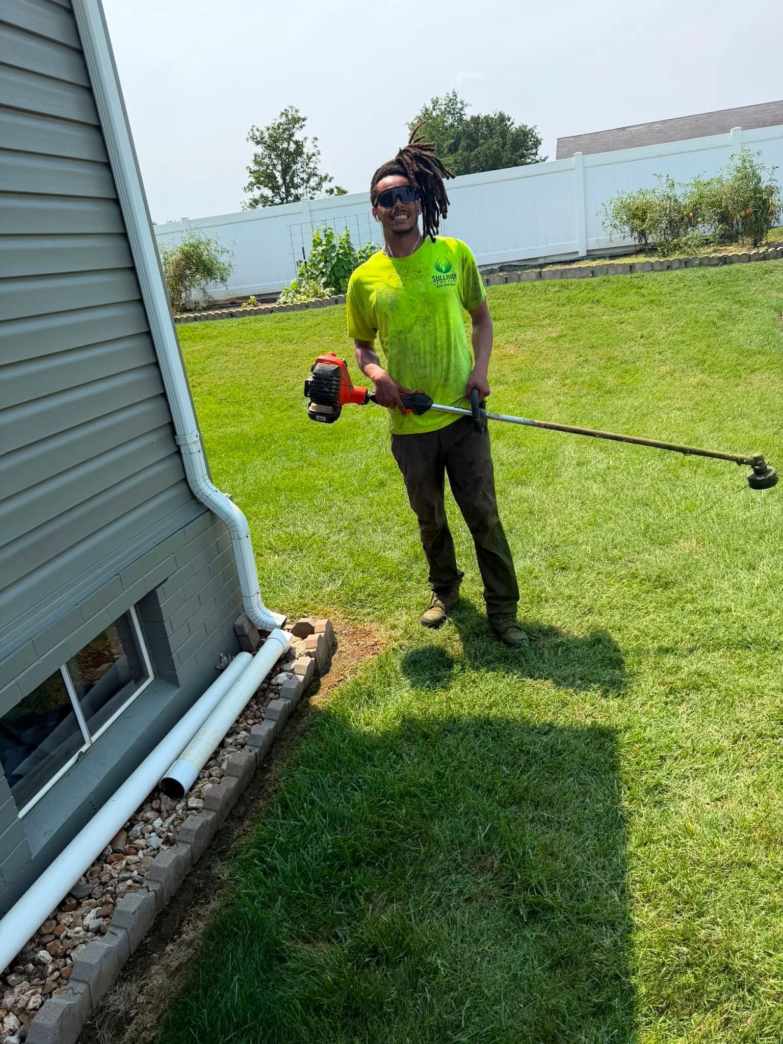 Person standing on grass beside a house, holding a string trimmer in bright sunlight