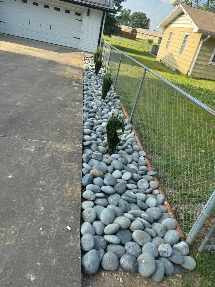 Stone-lined driveway beside a chain-link fence and grassy yard, with a garage in the background.