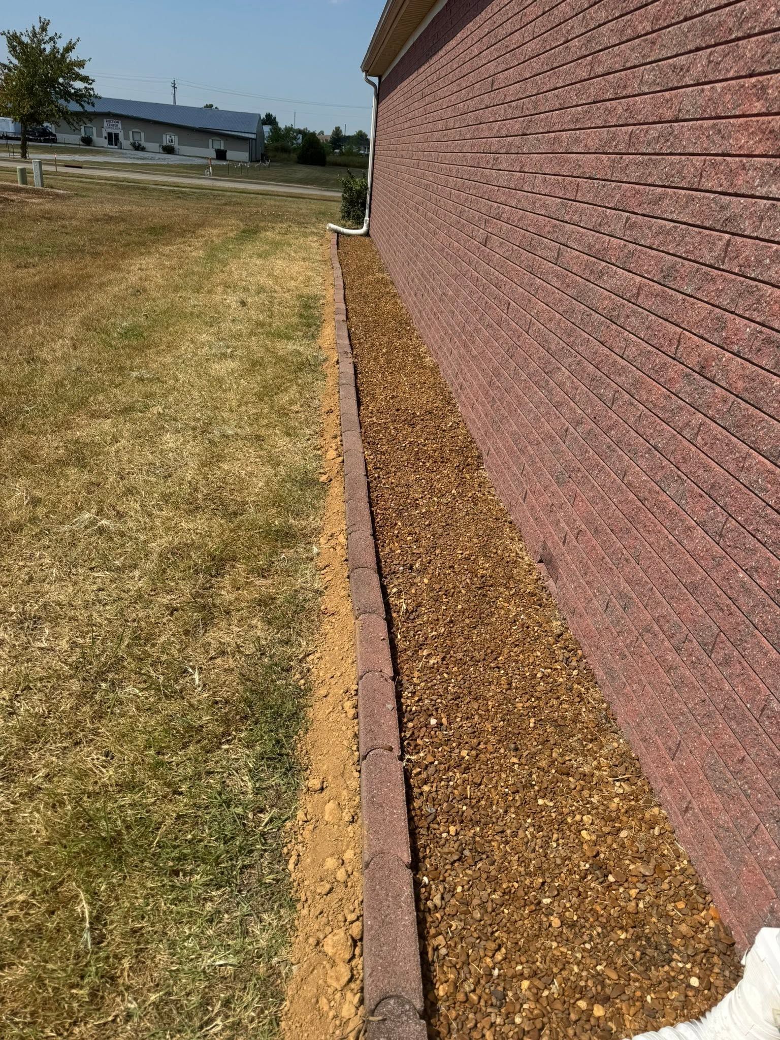 Narrow mulch bed runs along a brick wall beside a dry grassy field under a clear sky