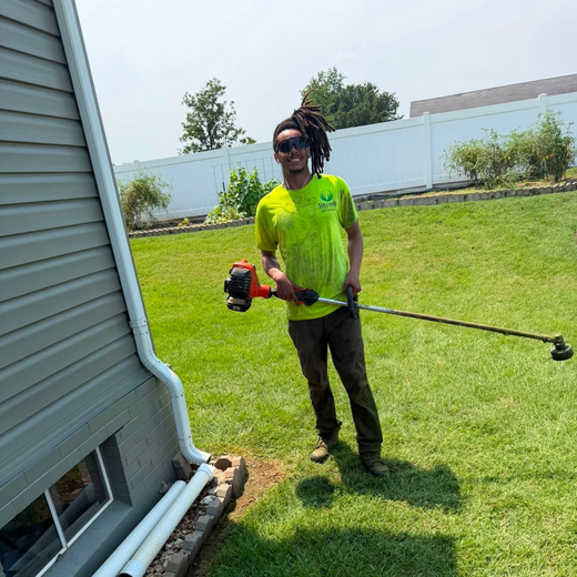 Person using a string trimmer on a sunny lawn beside a house and white fence