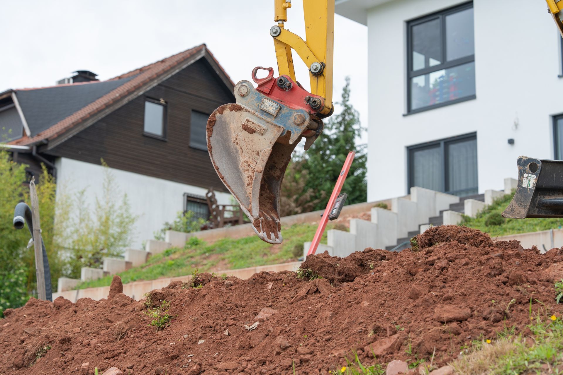 Excavator bucket digging red soil beside a house and steps on a sloped yard